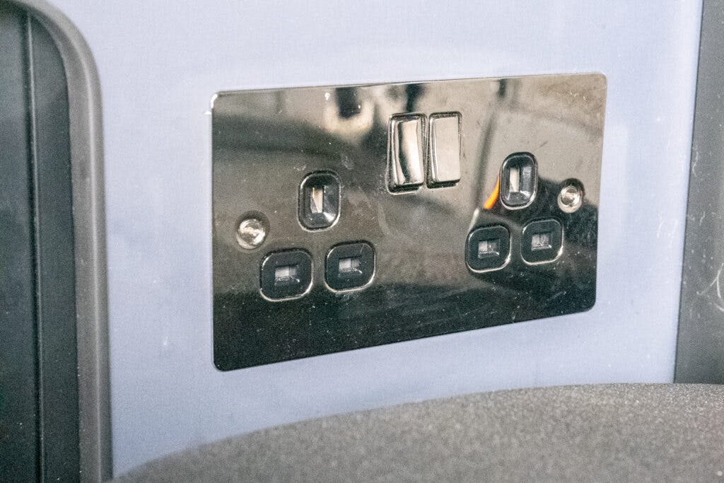 A metallic electrical outlet with a sleek design featuring four rectangular sockets and two switches is mounted on a light-colored wall, reminiscent of the modern amenities found in a 2021 Ford Transit Custom Camper. Two screws are visible on the left and right sides of the outlet plate.