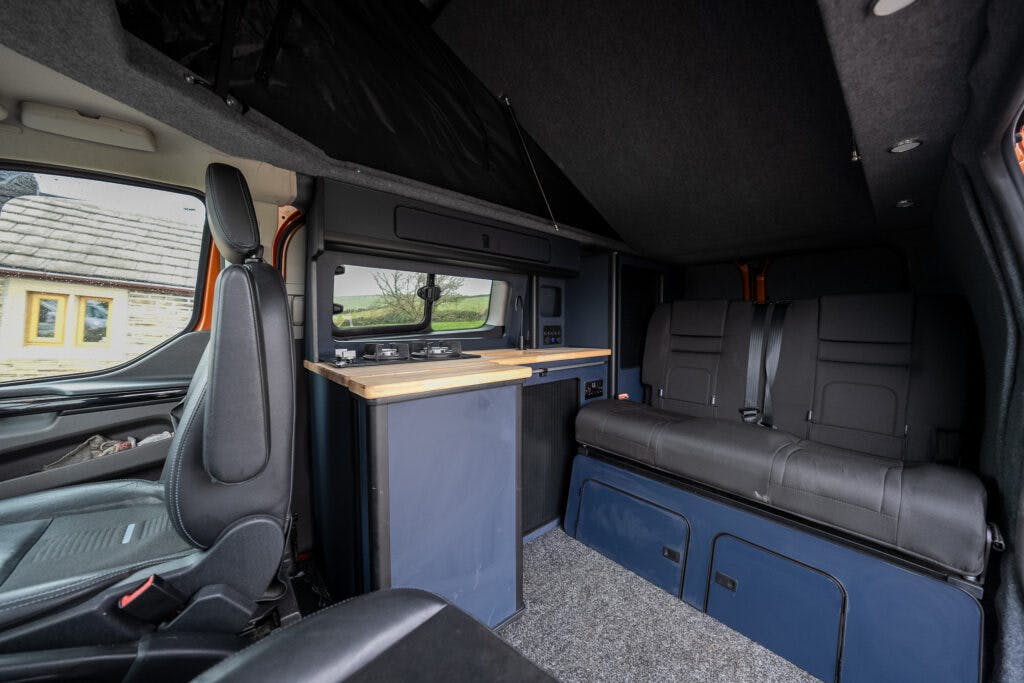 Interior view of the 2021 Ford Transit Custom Camper featuring two black seats, a small kitchen area with a wooden countertop and sink, storage compartments, and large windows providing natural light. The space is compact and designed for functional living.