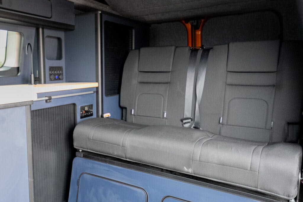 The image shows the interior of a 2021 Ford Transit Custom Camper. A folded-down grey seat with seat belts rests against a dark blue background. On the left, closed cabinets and a control panel are visible, with a light wooden countertop above them.