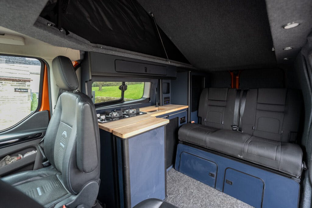 Interior view of a 2021 Ford Transit Custom Camper featuring a kitchenette with stove and sink on the left and a seating area with black cushioned seats on the right. The van interior is designed with functional storage compartments and a cozy atmosphere.