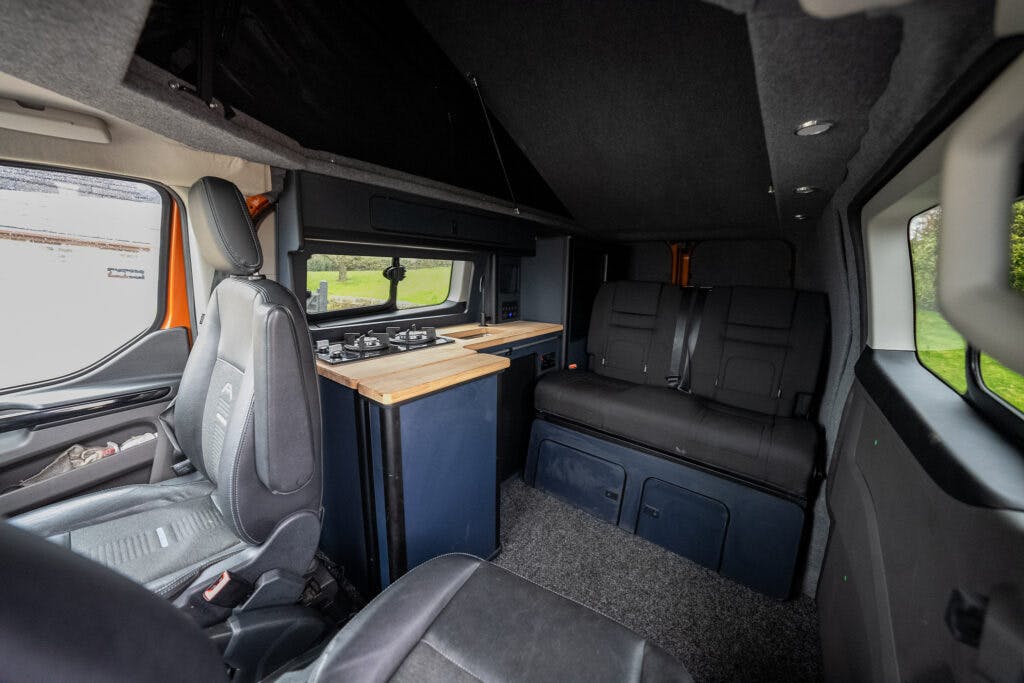 The image shows the interior of a 2021 Ford Transit Custom Camper. It features a driver and passenger seat, a countertop with a built-in stove, a seating area with black upholstery, overhead storage, and side windows. The interior is designed for compact living space.