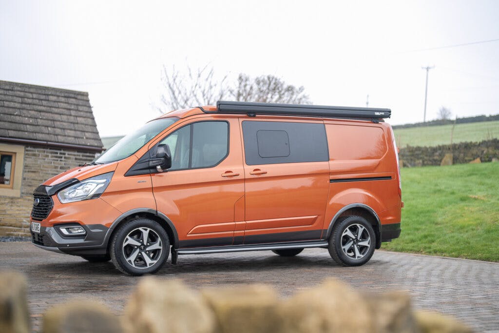 A brightly colored orange 2021 Ford Transit Custom Camper is parked on a paved driveway. The van has black trim and decals, with a roof rack attached. In the background, there is a stone building on one side and green grass with a stone wall on the other side. The sky appears overcast.