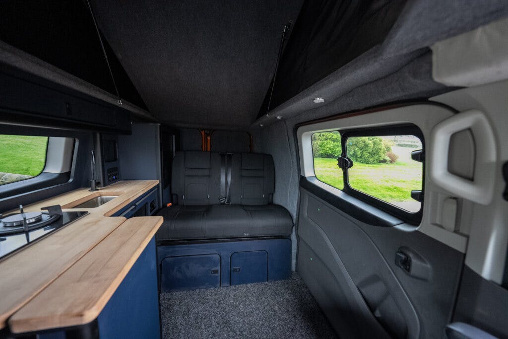 The interior of the 2021 Ford Transit Custom Camper features a compact kitchenette with a stovetop and wooden countertops on the left, black bench seating that appears to convert into a bed in the center, and large windows on the right, offering a view of the greenery outside.