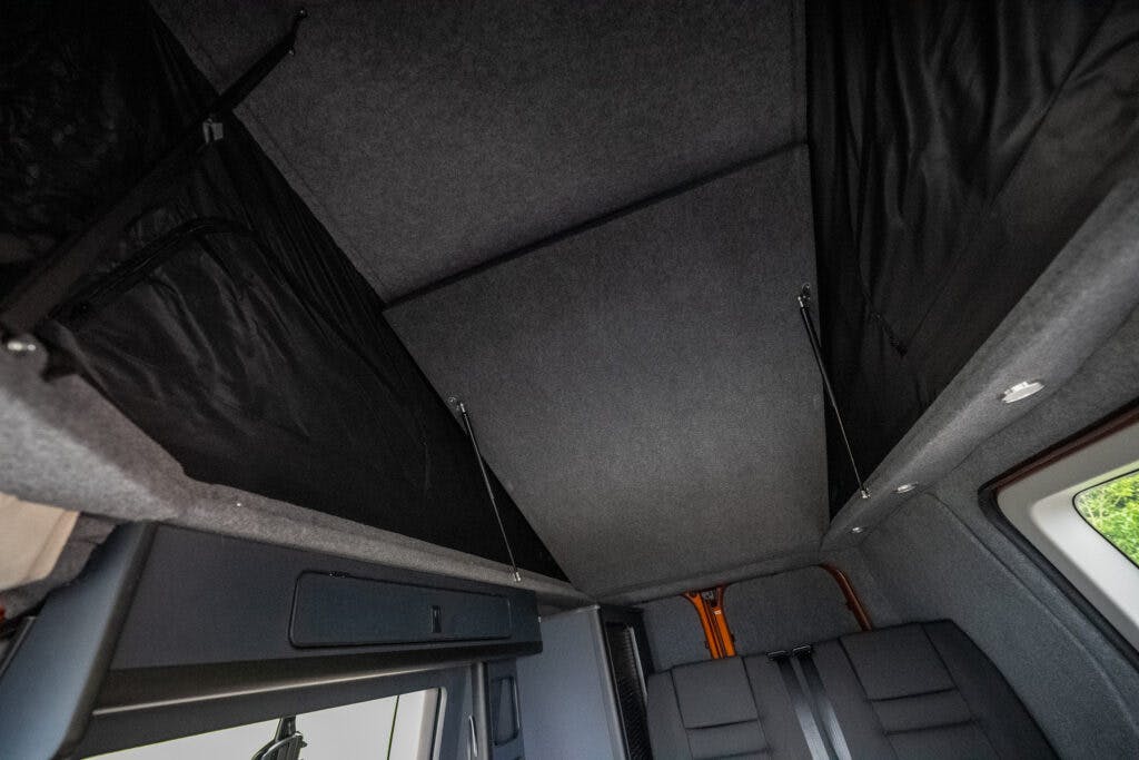 Interior view of a 2021 Ford Transit Custom Camper's roof displaying two raised, black fabric storage compartments. The surroundings are lined with dark gray upholstery, and parts of the seating area are visible at the bottom of the image.