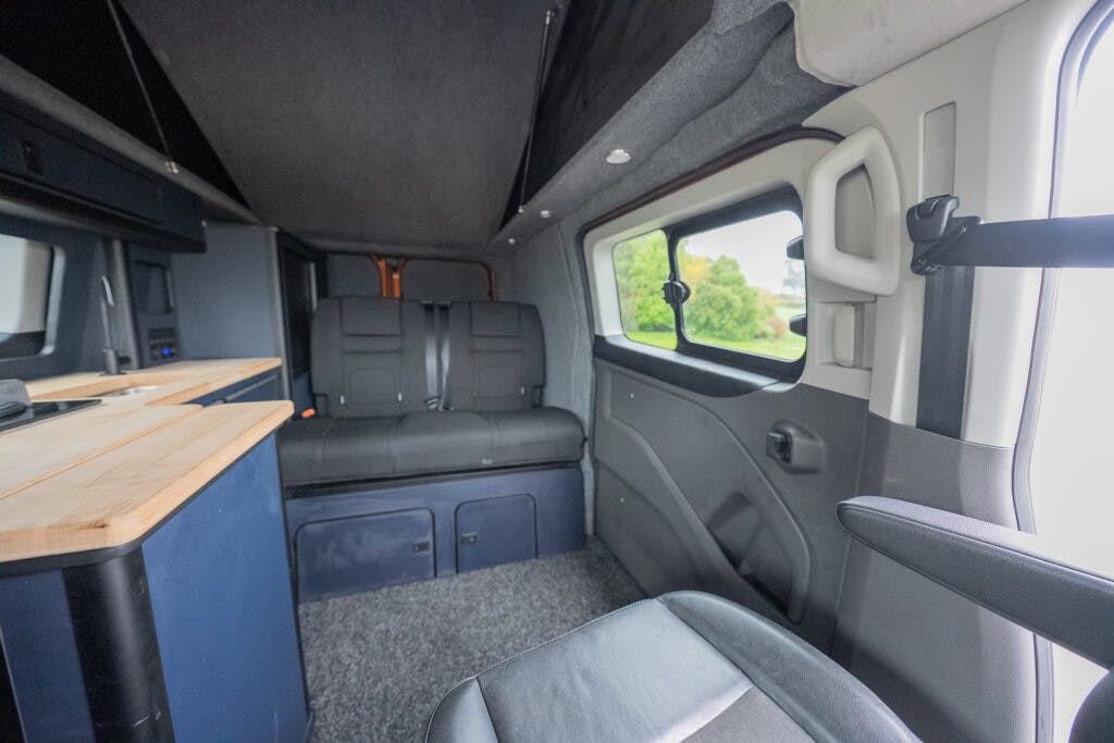 The interior of the 2021 Ford Transit Custom Camper features black seats, a wooden countertop, and large windows on the side. The seating area offers multiple reclining seats and ample space. The floor is carpeted, and the vehicle walls boast elegant gray and white detailing.