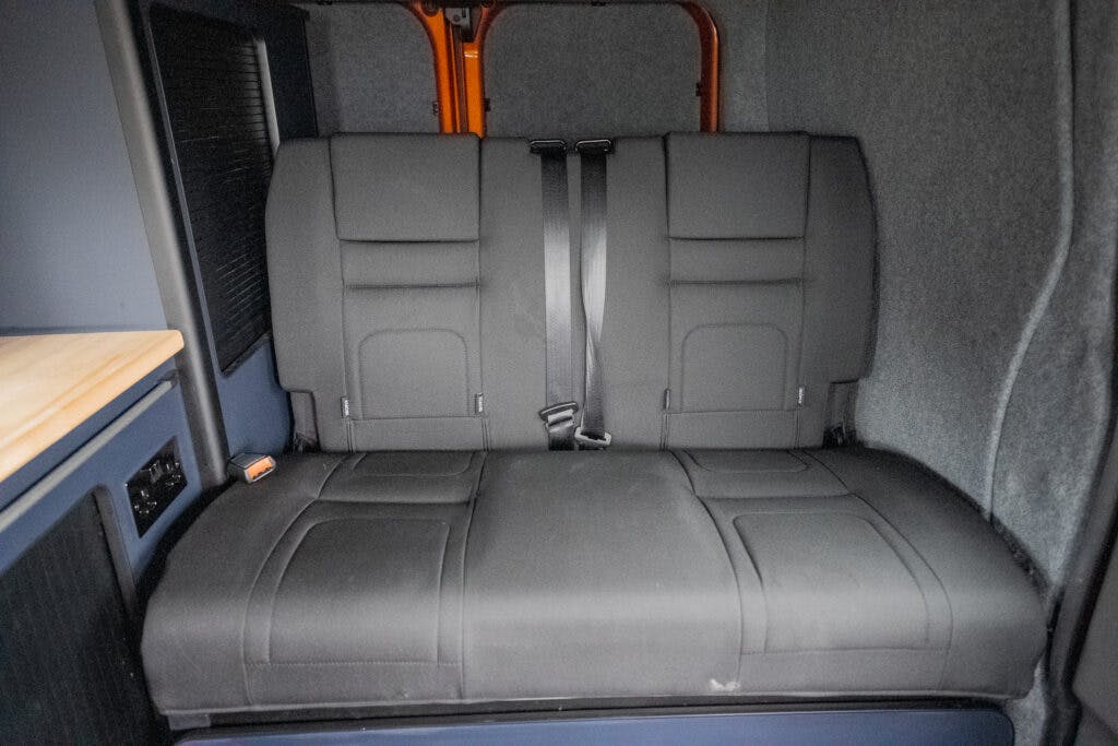 The image shows the rear interior of a 2021 Ford Transit Custom Camper, featuring a gray three-seater bench with two black seat belts. The bench is surrounded by dark panels and carpeting. Part of a wooden counter is visible on the left.
