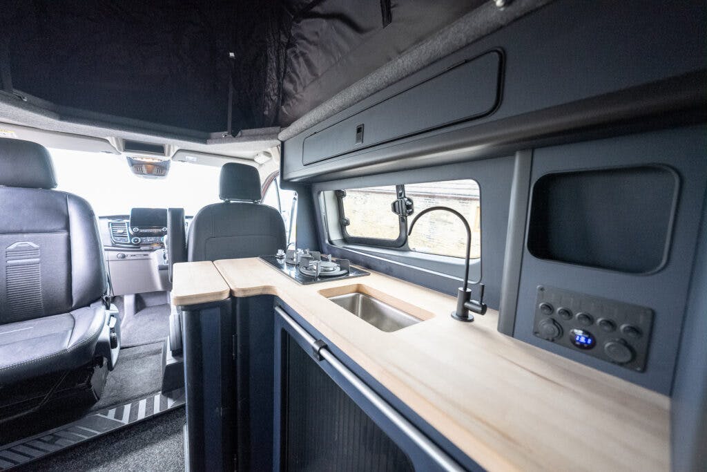 The image shows the interior of a 2021 Ford Transit Custom Camper. It features a compact kitchen with a wooden countertop, sink, and stove. The driver and passenger seats are visible, along with overhead storage compartments and a control panel.