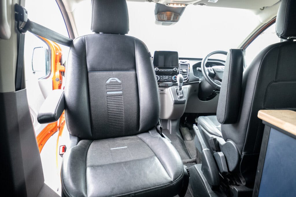 The interior of a 2021 Ford Transit Custom Camper showcases black leather front seats, a dashboard with a touchscreen display, steering wheel, and gearshift. The driver's side door is open, revealing an orange exterior.