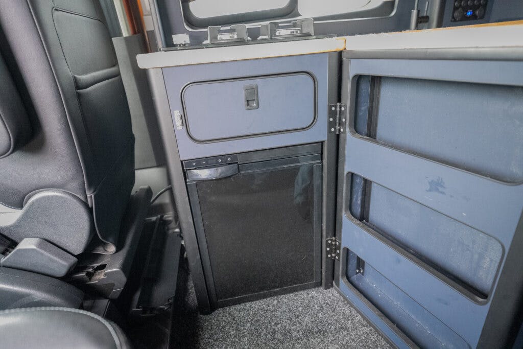 An interior view of a 2021 Ford Transit Custom Camper reveals a small black refrigerator installed beneath a blue cabinet with drawers. The area adjacent to the driver's seat is carpeted, and various control buttons are visible on the panel above the cabinet.