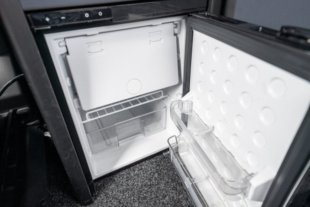 Image of an open mini refrigerator. The fridge has a small freezer compartment at the top and an empty shelf below. The door features a transparent, narrow shelf. The interior is clean and mostly empty. Set against the backdrop of a cozy 2021 Ford Transit Custom Camper with its stylish black exterior on carpeted flooring.