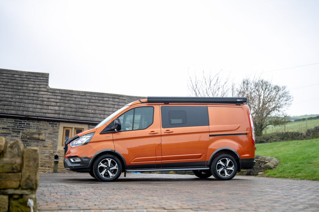 A bright orange 2021 Ford Transit Custom Camper with a black retractable awning is parked on a cobblestone driveway in front of a stone building with a tiled roof. The background features a grassy lawn and some bare trees, indicating it's fall or winter.