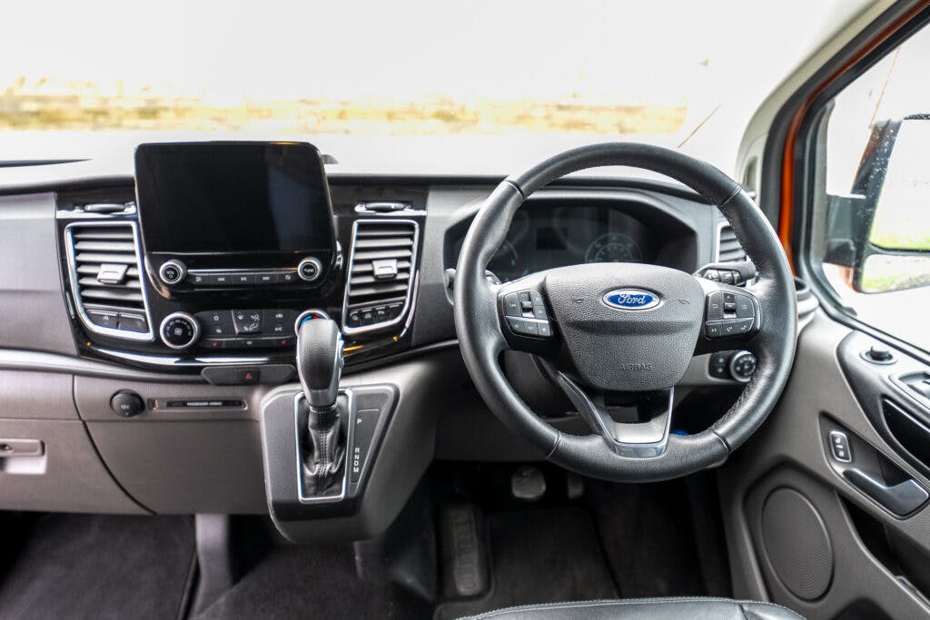 The image shows the interior of a 2021 Ford Transit Custom Camper, focusing on the driver's area. It includes a steering wheel with control buttons, a central touchscreen display, various control knobs and buttons, and a gear shift lever.