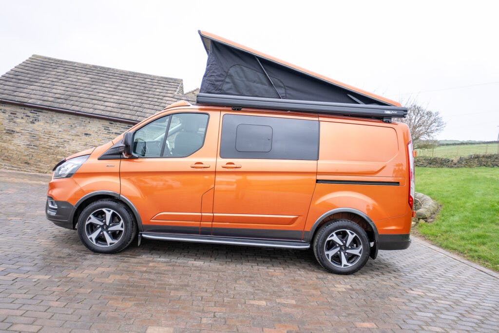 A 2021 Ford Transit Custom Camper with a pop-up roof is parked on a cobblestone driveway. The orange van features black accents and alloy wheels. The pop-up roof is partially extended, with a stone building and green lawn in the background.