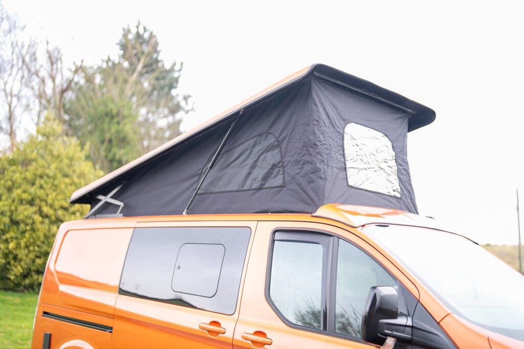 A bright orange 2021 Ford Transit Custom Camper van with a pop-up roof tent partially extended. The tent is black with a reflective window covering, allowing additional space and light into the van. The background shows a cloudy sky, green trees, and shrubs.