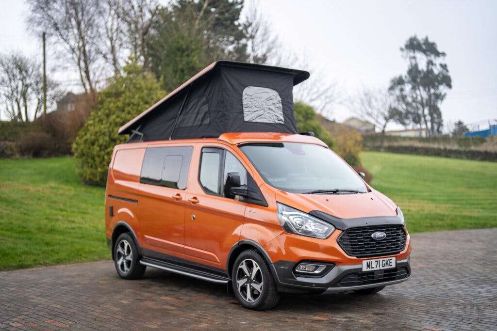 A 2021 Ford Transit Custom Camper with a pop-up roof is parked on a paved area. The bright orange van features tinted windows and sleek black accents. The extended roof reveals a black fabric section, while trees and grassy areas create a serene backdrop under an overcast sky.