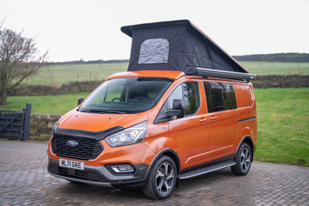 An orange 2021 Ford Transit Custom Camper with a pop-up roof is parked on a cobblestone driveway. The license plate reads "ML71 GKE." The surrounding area features a green field and trees, with a wooden fence visible in the background.