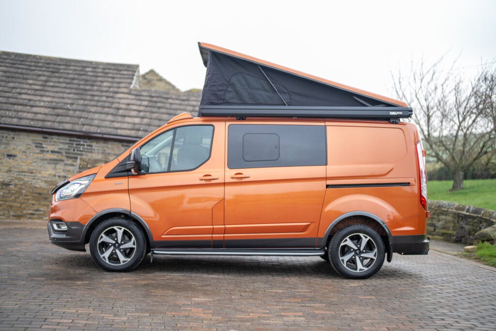 A side view of a 2021 Ford Transit Custom Camper with a pop-up roof, parked on a cobblestone driveway. The pop-up roof is extended, and the van has black windows and alloy wheels. The background includes a stone building and leafless trees.
