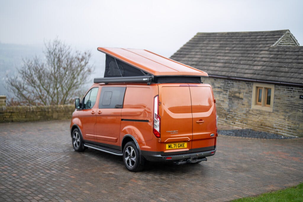 A 2021 Ford Transit Custom Camper, painted a vibrant orange, is parked on a stone-paved driveway next to a stone house. The van's pop-up roof is extended. Its license plate reads "ML71 GWE." The background features leafless trees and a foggy sky, suggesting a possibly cold day.