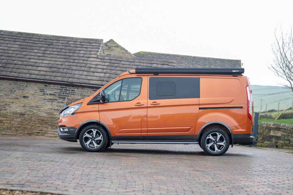A side view of a 2021 Ford Transit Custom Camper in vibrant orange with tinted windows parked on a brick pavement. The vehicle has a black roof rack and alloy wheels. The background features a stone building with a sloped roof and a partial view of countryside scenery.