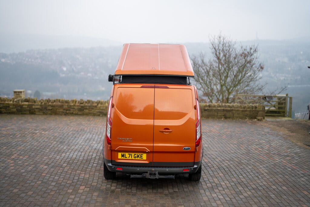 A bright orange 2021 Ford Transit Custom Camper with a raised roof is parked on a cobblestone surface. The rear of the van is visible, including a license plate with the number "ML71 GKE." The background features a foggy, hilly landscape and a leafless tree.