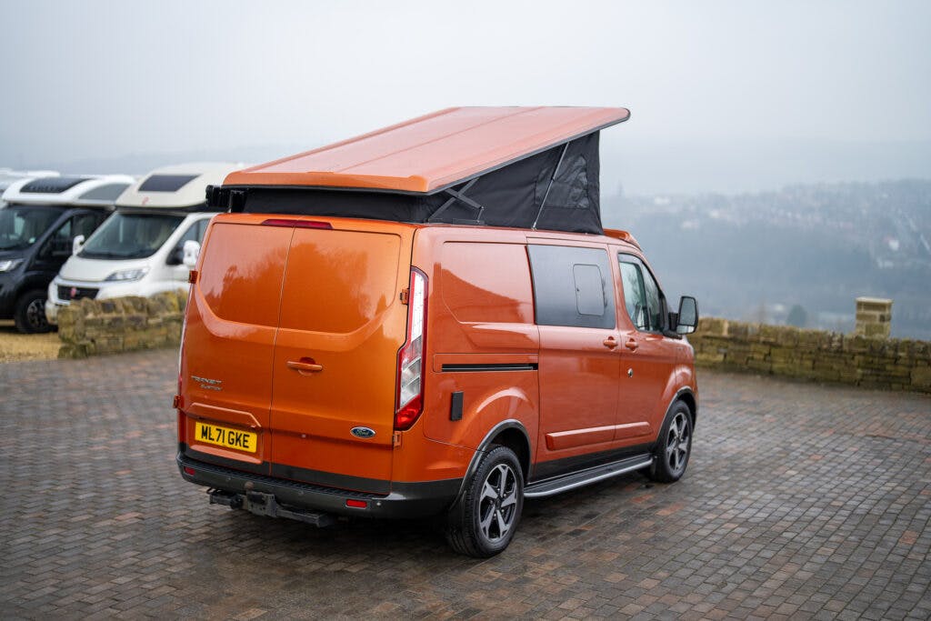 An orange 2021 Ford Transit Custom Camper with an elevated pop-up roof is parked on a paved area overlooking a foggy landscape. The vehicle has a UK license plate reading "ML17 GKE." Several other camper vans are visible in the background.