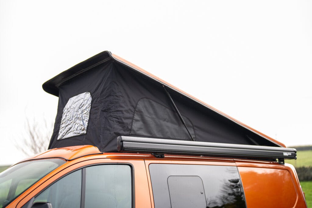 The image shows an orange 2021 Ford Transit Custom Camper featuring a pop-up roof tent. The tent is black with a fabric window and appears to be extended, increasing the vertical space within the vehicle. An awning is also attached to the side of the van. The background is blurred and primarily white.