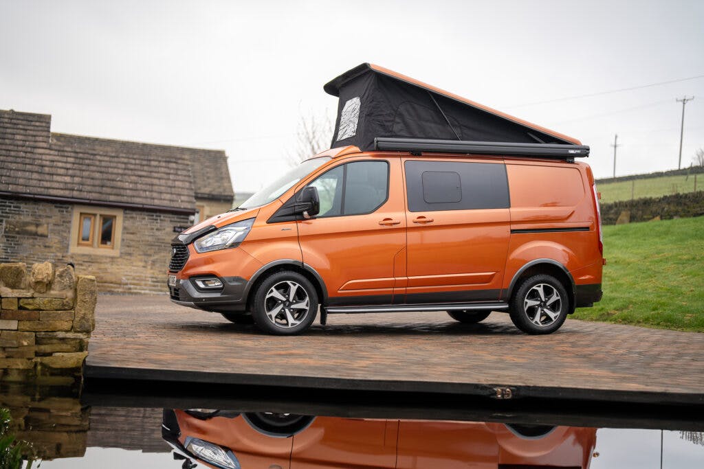 A 2021 Ford Transit Custom Camper with a pop-up roof is parked on a cobblestone driveway near a stone building. The surrounding area features a grassy landscape and a small body of water reflecting the van.
