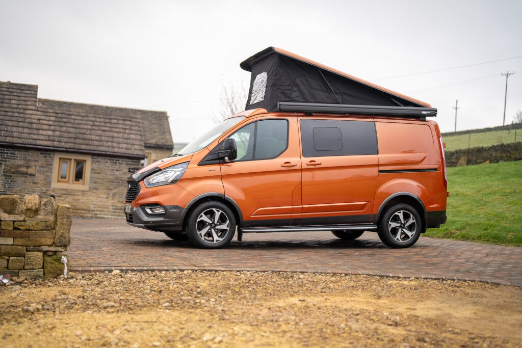 A 2021 Ford Transit Custom Camper in a vibrant orange hue is parked on a brick driveway beside a stone house. The camper van boasts an extended pop-top roof, enhancing its versatility. Surrounding the scene are a stone wall and lush grassy landscape, all under an overcast sky.