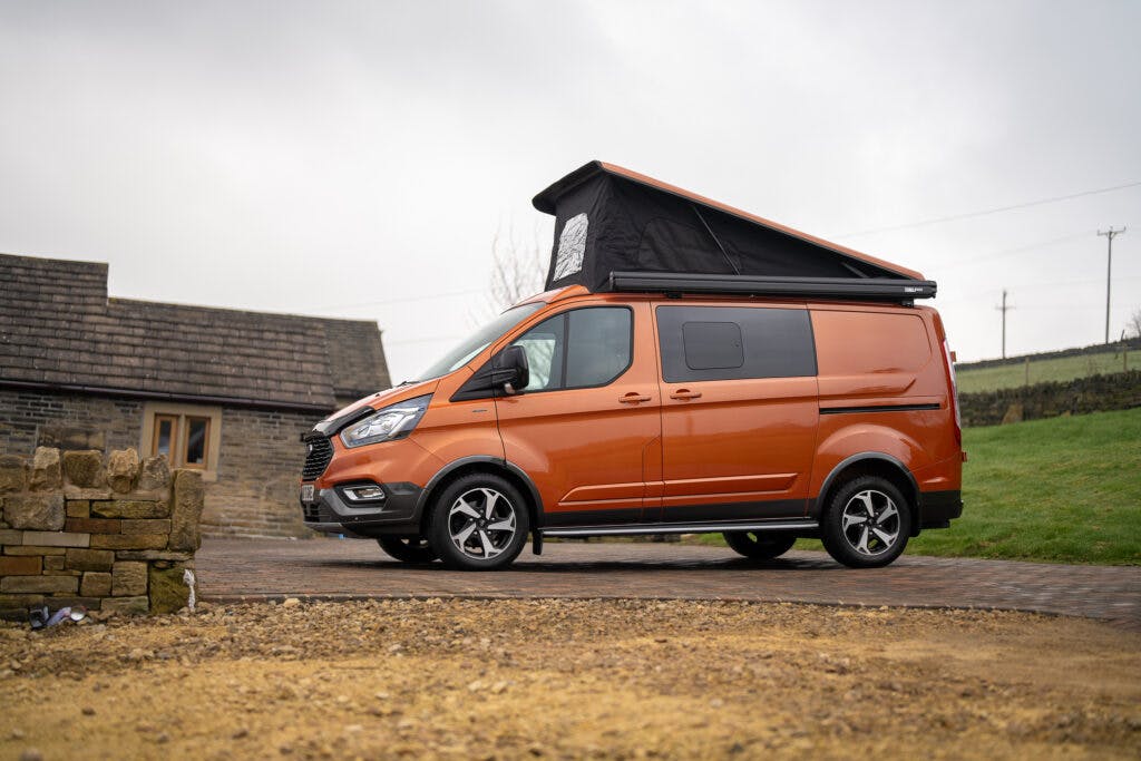 A bright orange 2021 Ford Transit Custom Camper with an extended black roof is parked on a paved driveway. The background includes a stone building and a grassy field under an overcast sky.