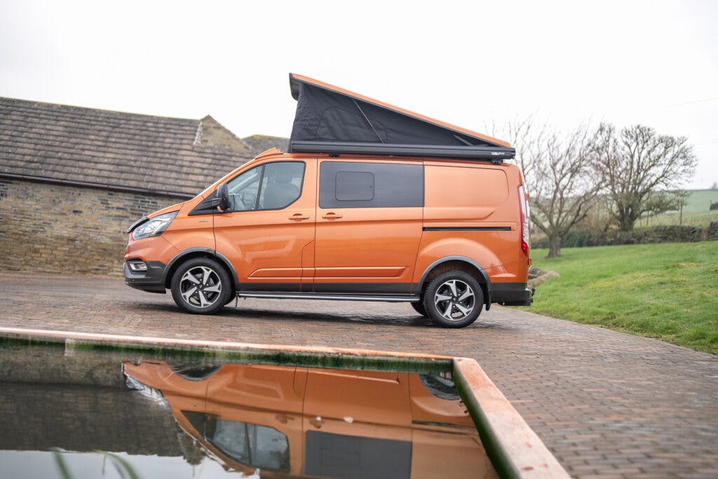 A 2021 Ford Transit Custom Camper with a partially elevated roof is parked on a paved area near a reflective water surface. The background shows a rustic stone building and a grassy area with some trees.