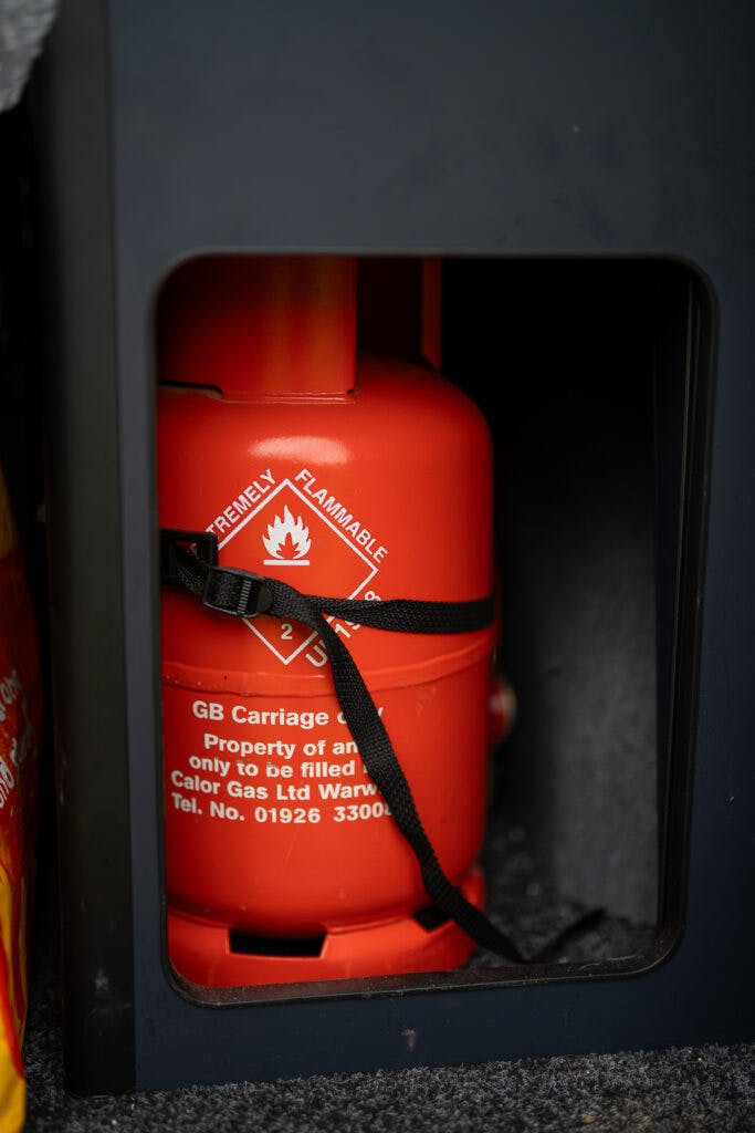 A close-up of a red gas cylinder strapped inside a 2021 Ford Transit Custom Camper compartment. The cylinder has "Extremely Flammable" with a flame symbol and the label "GB Carriage - Property of Calor Gas Ltd." A phone number and other text details are visible.