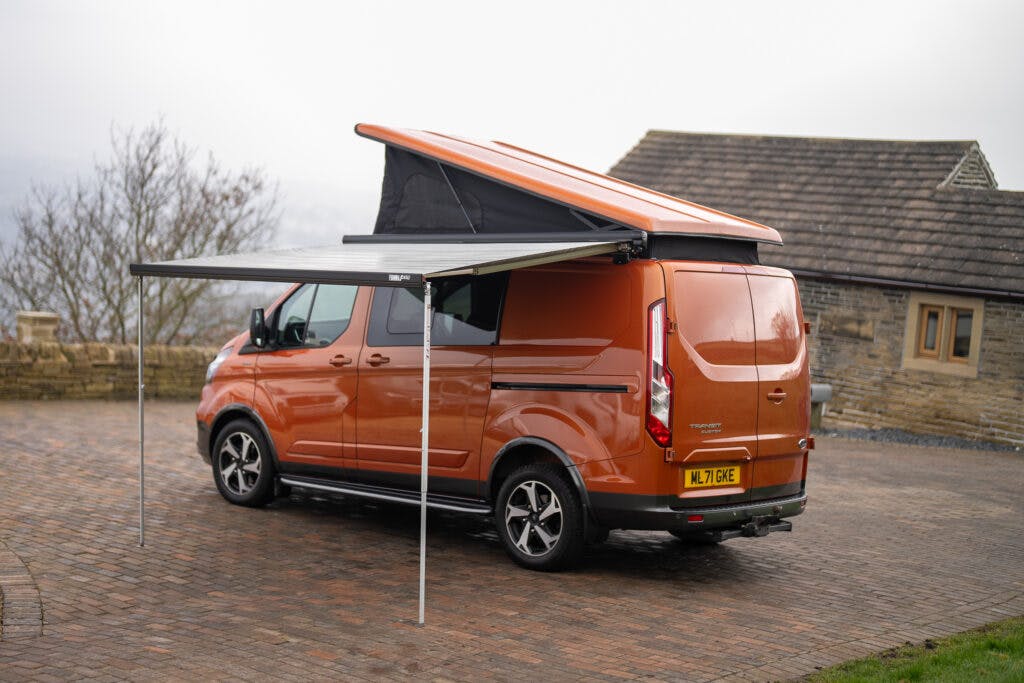 A 2021 Ford Transit Custom Camper is parked on a brick driveway with its rooftop tent popped open and an awning extended on the side. A stone house with a small window is in the background, and leafless trees are partially visible on the left.