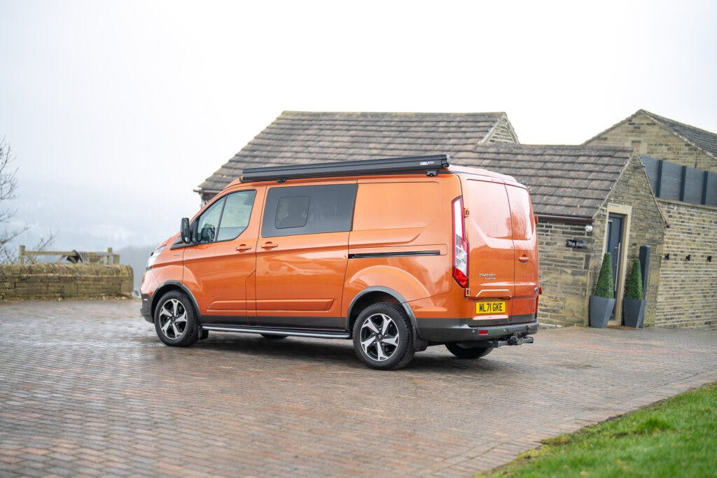 A parked orange 2021 Ford Transit Custom Camper with a Western Europe license plate faces away from the camera. The van has tinted windows and a side step. In the background, there is a stone building with a tiled roof and a foggy landscape.