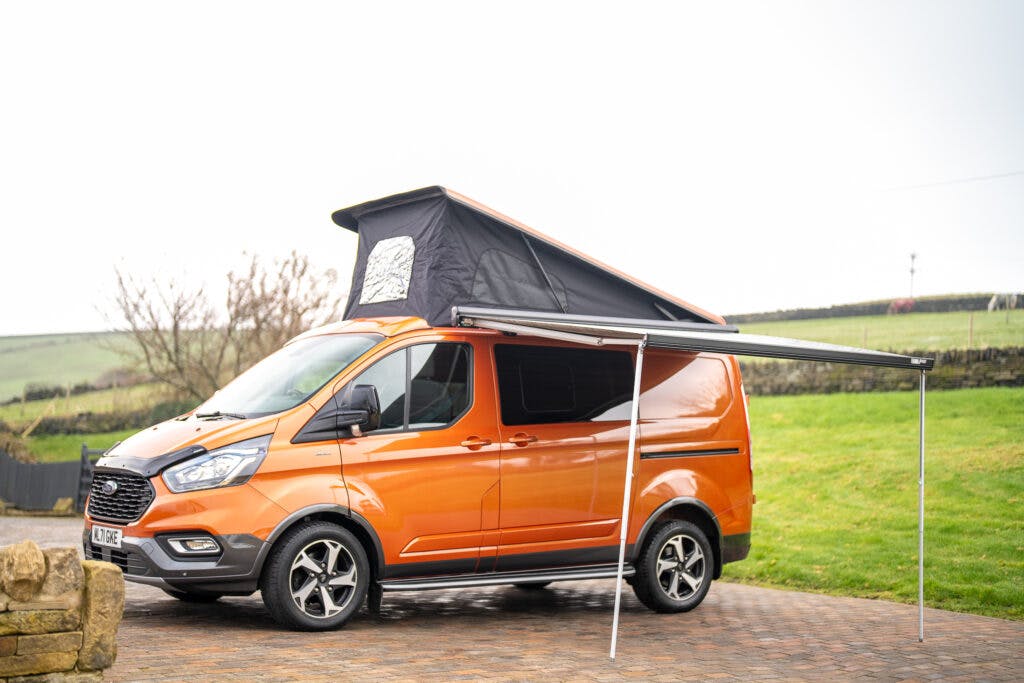 A 2021 Ford Transit Custom Camper in vibrant orange is parked in a driveway, its roof tent extended upwards and an awning pulled out. The backdrop consists of lush green fields and a cloudy sky.