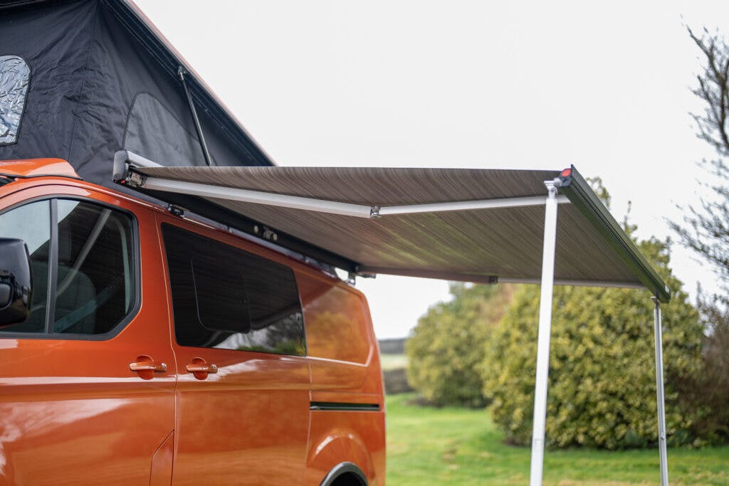 An orange 2021 Ford Transit Custom Camper van is parked with its side awning extended. The awning provides shade and extends from the roof of the van, supported by poles. A pop-up tent is visible on the roof of the van, and the background features greenery and bushes.