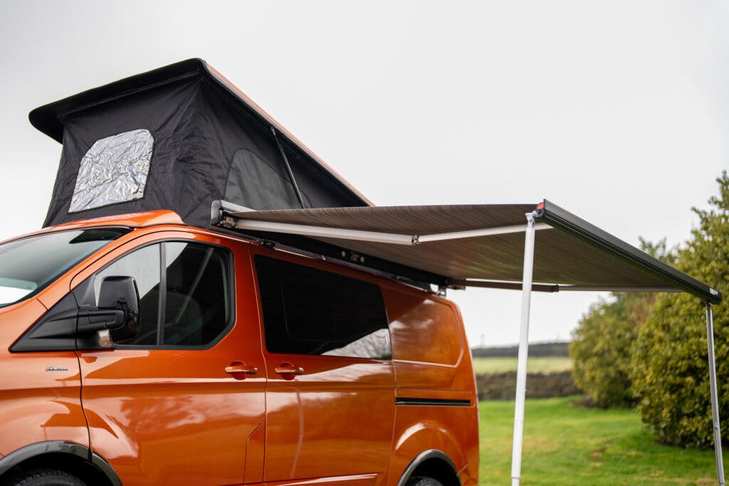 A bright orange 2021 Ford Transit Custom Camper is shown with a pop-up roof extended and an awning deployed. The scene takes place outdoors on a grassy area with overcast skies, and the campervan appears to be set up for camping.