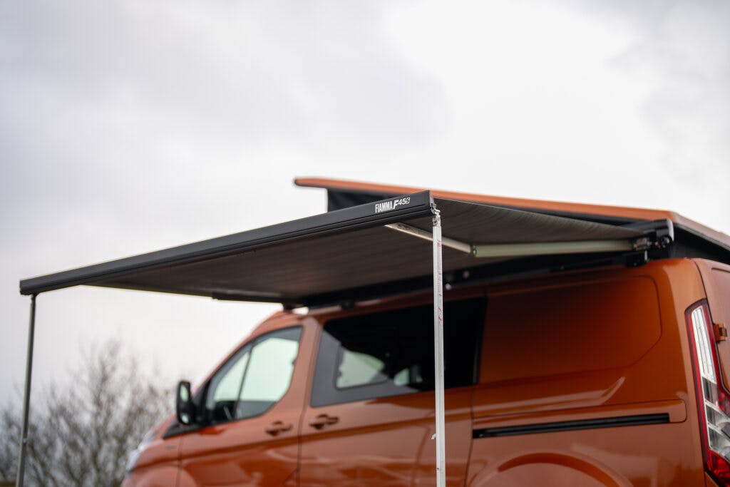 An orange 2021 Ford Transit Custom Camper with a black roof awning supported by a white pole extends out, providing shade. The awning has the logo "Front Runner" visible on the corner. The sky in the background is mostly cloudy.