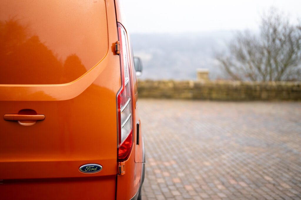 A close-up view of the back of an orange 2021 Ford Transit Custom Camper, parked on a brick-paved surface. The left edge of the van, including the rear light and Ford emblem, is visible. The background features a misty landscape with a stone wall and bare trees.