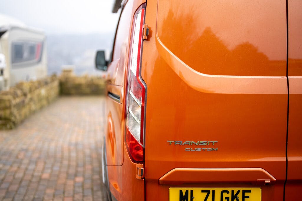 An orange 2021 Ford Transit Custom Camper van is parked on a cobblestone driveway. The rear of the van is shown prominently, with the "Transit Custom" badge and a yellow license plate in view. Other vehicles and a stone wall are visible in the background.