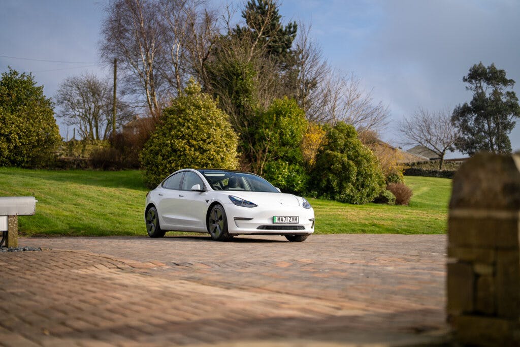 A white 2021 Tesla Model 3 Long Range AWD is parked on a brick driveway with a well-maintained lawn and trees in the background. The sky is partly cloudy, and a license plate is visible at the front of the car.