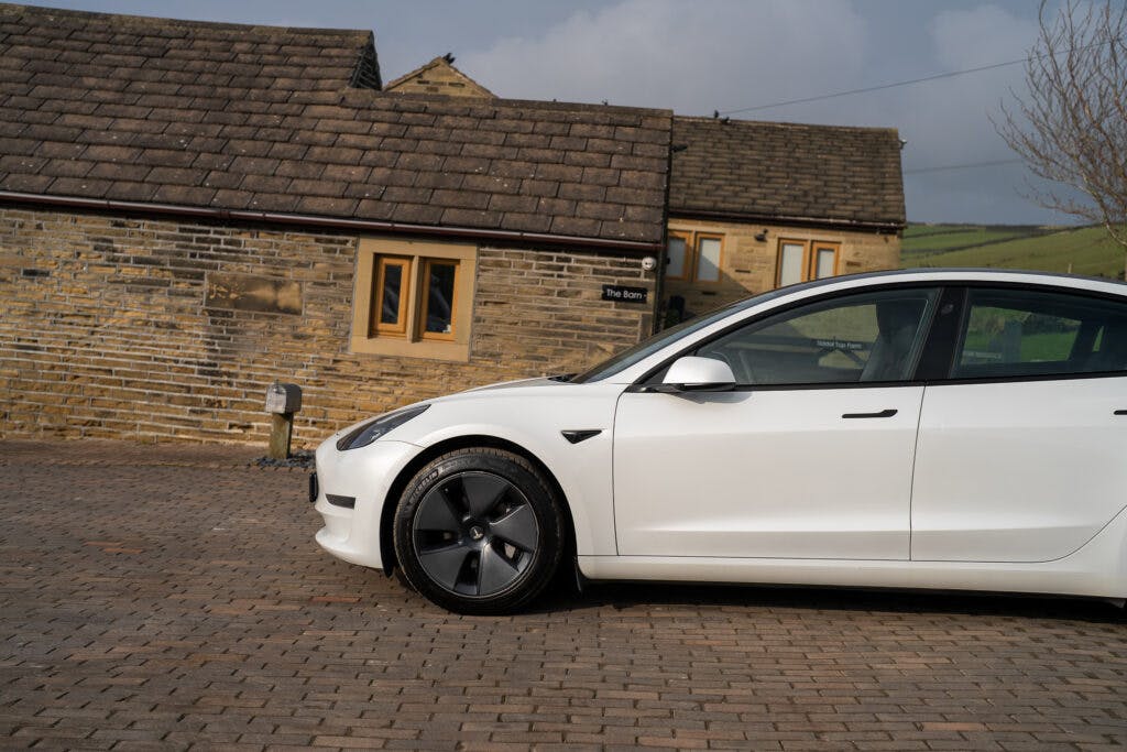 A 2021 Tesla Model 3 Long Range AWD is parked on a cobblestone driveway in front of a stone house with a pitched roof. The house has two windows visible, and part of a green hillside is in the background. The sky is partly cloudy.