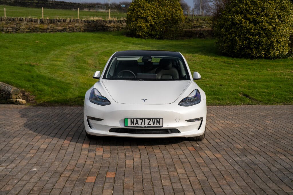 A white 2021 Tesla Model 3 Long Range AWD is parked on a brick-paved driveway. The car's front, including the license plate "MA71 ZVM," is visible. The background displays a grassy area, bushes, and a partially visible stone wall.