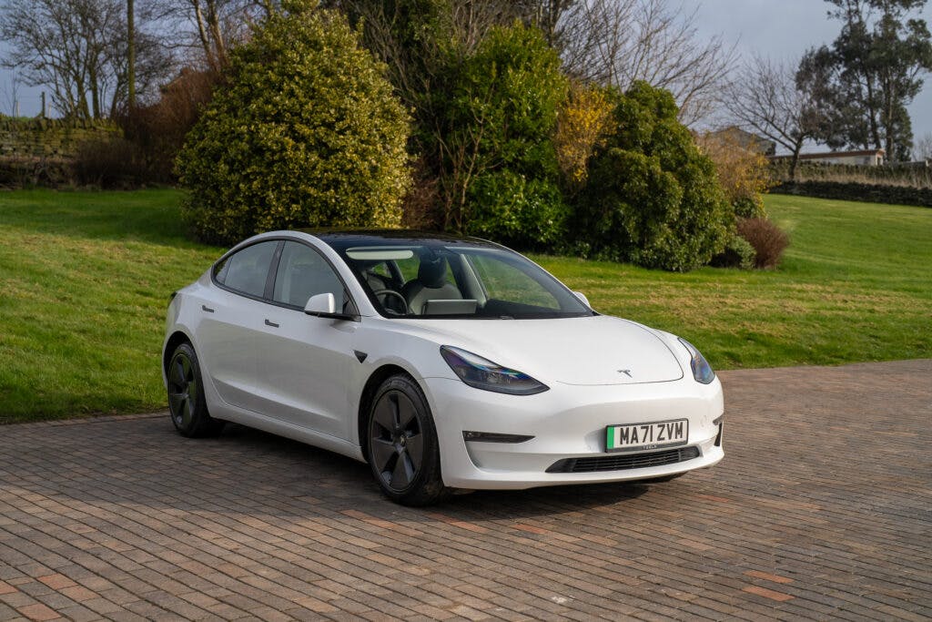A white 2021 Tesla Model 3 Long Range AWD is parked on a brick driveway in front of a grassy area with shrubs and trees. The car's front and side are visible, sporting a license plate that reads "MA71 ZVW". The scene is outdoors during the daytime.