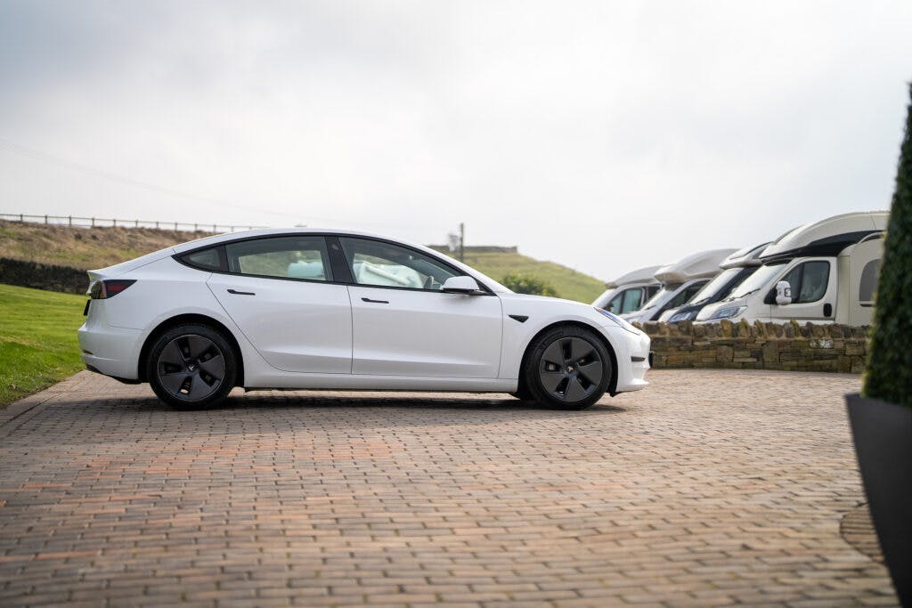 A white 2021 Tesla Model 3 Long Range AWD sedan is parked on a brick driveway. Several larger vehicles, including motorhomes, are lined up in the background. The sky is overcast, and the area appears to be a paved lot adjacent to a grassy field and stone wall.