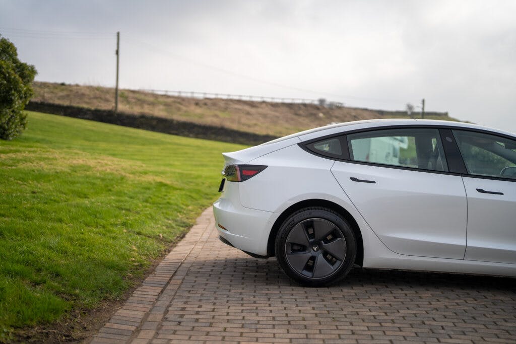 A white 2021 Tesla Model 3 Long Range AWD is parked on a brick-paved driveway next to a grassy area. The photo focuses on the rear half of the vehicle, highlighting the back wheel and taillight. A hill with a fence on top is visible in the background.