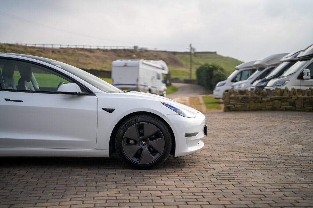 A 2021 Tesla Model 3 Long Range AWD is parked on a cobblestone driveway. In the background, several RVs are lined up alongside a grassy hill and a stone wall. The scene is outdoors on a cloudy day.