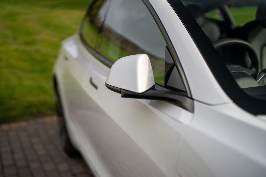 Close-up of a 2021 Tesla Model 3 Long Range AWD's side mirror and part of the front window. The white car is parked on a paved surface, with green grass visible in the background. The angle emphasizes the sleek and modern design of the vehicle.
