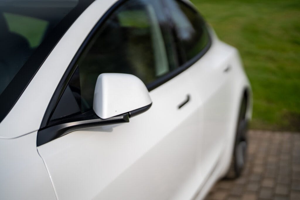 Close-up of the passenger side of a white 2021 Tesla Model 3 Long Range AWD, showing the sleek side mirror and part of the front door. The car is parked on a paved area with vibrant green grass in the background.