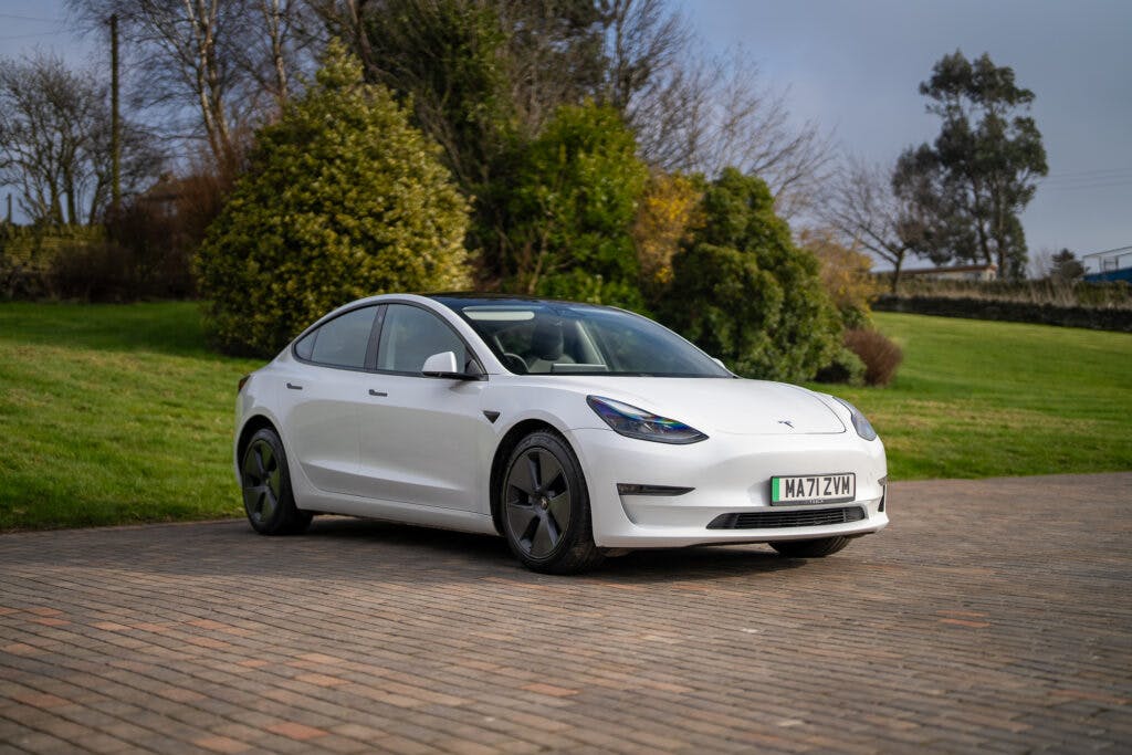 A white 2021 Tesla Model 3 Long Range AWD is parked on a paved area with a grassy, tree-lined background. The car is facing slightly to the right. The surroundings include shrubs, trees, and a partly cloudy sky. The license plate reads "MA72 YWM".