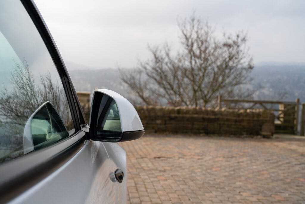 Close-up of the side mirror of a white 2021 Tesla Model 3 Long Range AWD parked on a cobblestone surface. In the background, a blurry view of bare trees and a stone wall, with an overcast sky and a faint glimpse of distant hills.